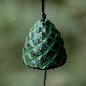 May include: A green wind chime bell with a textured, pinecone-like design. The bell is suspended by a black cord against a blurred green background. The bell's surface has dark accents.