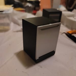 May include: A miniature dishwasher model featuring a matte black exterior casing and a light gray front panel with a horizontal handle. The appliance has a rectangular, boxy shape and is designed as a small-scale replica of a kitchen unit.