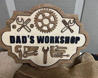 May include: Wooden sign with the words "DAD'S WORKSHOP" in black letters. The sign features a gear, wrenches, and other tools. The sign is made of light and dark brown wood.