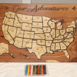 Może przedstawiać: Drewniana mapa Stanów Zjednoczonych z wygrawerowanym napisem "Our Adventures" u góry. Mapa jest podzielona na poszczególne stany i obejmuje Alaskę i Hawaje. W pierwszym planie znajduje się zestaw kolorowych ołówków.