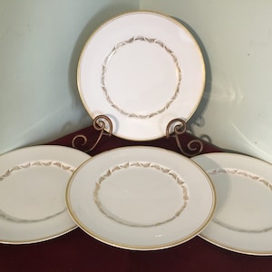 May include: Four white ceramic dinner plates with a gold rim and a delicate gold leaf design around the edge.