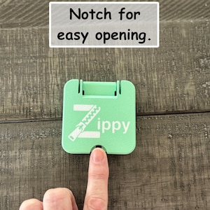 May include: A square, mint green plastic tool featuring a white graphic of a zipper and the text Zippy. The device includes a small notch at the bottom edge for easy opening, as indicated by the text Notch for easy opening above the object.