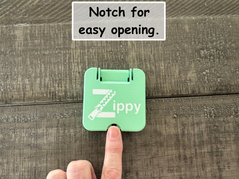 May include: A square, mint green plastic tool featuring a white graphic of a zipper and the text Zippy. The device includes a small notch at the bottom edge for easy opening, as indicated by the text Notch for easy opening above the object.