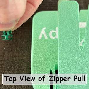 May include: A small green plastic zipper pull component held between fingers, positioned next to a larger mint green plastic tool with a textured surface. The text "Top View of Zipper Pull" is printed in a white rectangular box at the bottom.