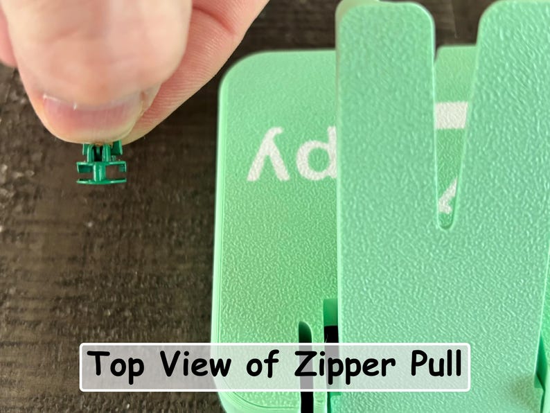 May include: A small green plastic zipper pull component held between fingers, positioned next to a larger mint green plastic tool with a textured surface. The text "Top View of Zipper Pull" is printed in a white rectangular box at the bottom.