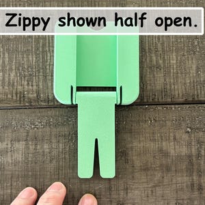 May include: A light green plastic Zippy tool shown in a half-open position. The device features a rectangular body with a hinged, bifurcated arm extending downward. The text "Zippy shown half open." is printed in a white box at the top of the image.