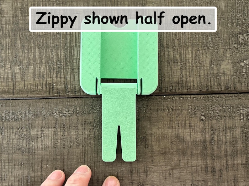 May include: A light green plastic Zippy tool shown in a half-open position. The device features a rectangular body with a hinged, bifurcated arm extending downward. The text "Zippy shown half open." is printed in a white box at the top of the image.