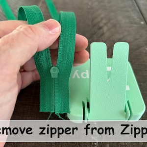 May include: A bright green zipper being held next to a light mint green plastic tool with a U-shaped slot. The tool features the text Zippy printed on its surface. A white rectangular label at the bottom contains the text Remove zipper from Zippy.