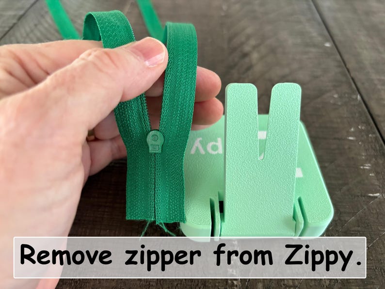 May include: A bright green zipper being held next to a light mint green plastic tool with a U-shaped slot. The tool features the text Zippy printed on its surface. A white rectangular label at the bottom contains the text Remove zipper from Zippy.