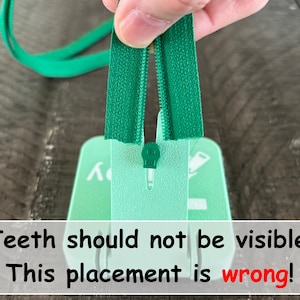 May include: A green zipper being inserted into a light green plastic tool. The text on the tool reads: Teeth should not be visible! This placement is wrong! The zipper tape is green with matching teeth, and the tool has a central slot for alignment.