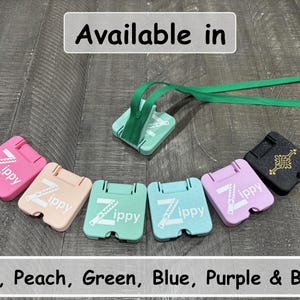 May include: A collection of square zipper pull aids in pink, peach, light green, blue, and purple, each featuring the white text Zippy and a zipper graphic. A black version features a gold Celtic knot design. One green pull includes a green ribbon.