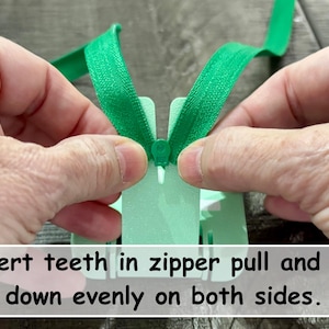 May include: A bright green zipper being threaded through a light mint green plastic zipper installation tool. The tool features a central slot for the zipper slider and includes the printed text: Insert teeth in zipper pull and pull down evenly on both sides.