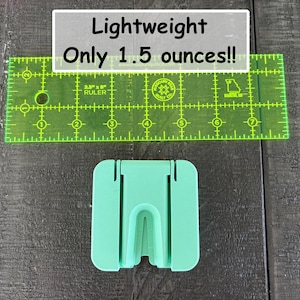 May include: A mint green plastic stand with a central U-shaped cutout, positioned below a transparent green 2.5-inch by 8-inch ruler. The text on the image reads: Lightweight Only 1.5 ounces!! The stand features a flat base and vertical slots.