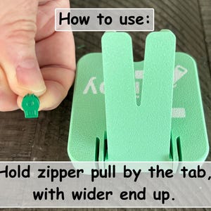 May include: A mint green plastic zipper aid tool with a V-shaped upright support. The tool features white printed text that reads How to use: and Hold zipper pull by the tab, with wider end up. A small green zipper pull is held near the tool.