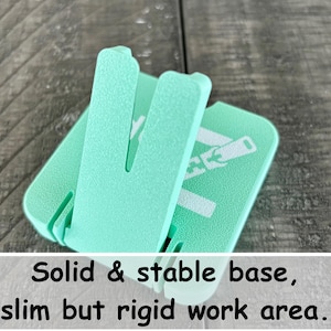 May include: A mint green plastic stand with a textured surface and a hinged support arm. The base features the printed text: Solid & stable base, slim but rigid work area. The object has a square-shaped footprint with rounded corners and a vertical slot.