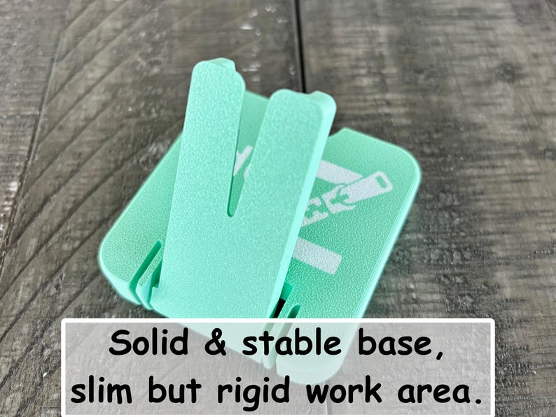 May include: A mint green plastic stand with a textured surface and a hinged support arm. The base features the printed text: Solid & stable base, slim but rigid work area. The object has a square-shaped footprint with rounded corners and a vertical slot.