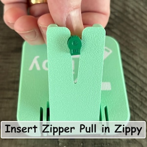 May include: A light mint green plastic tool with a vertical slot designed to hold a small green zipper pull. The device features a textured surface and white printed text that reads Insert Zipper Pull in Zippy, providing a guide for use.