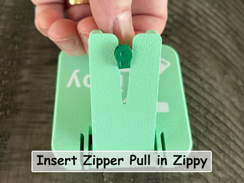May include: A light mint green plastic tool with a vertical slot designed to hold a small green zipper pull. The device features a textured surface and white printed text that reads Insert Zipper Pull in Zippy, providing a guide for use.