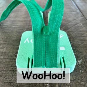 May include: A mint green square plastic base with a central vertical slot holding a matching green zipper tape and slider. The base features white printed text that reads WooHoo! with a small graphic icon visible on the top right corner surface.