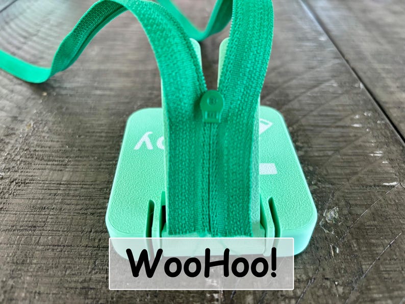 May include: A mint green square plastic base with a central vertical slot holding a matching green zipper tape and slider. The base features white printed text that reads WooHoo! with a small graphic icon visible on the top right corner surface.