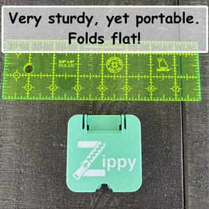 May include: A mint green square folding stand with the white text Zippy and a zipper graphic. A transparent neon green ruler is positioned above the stand, showing a width of approximately 2.5 inches. The text Very sturdy, yet portable. Folds flat! is visible.
