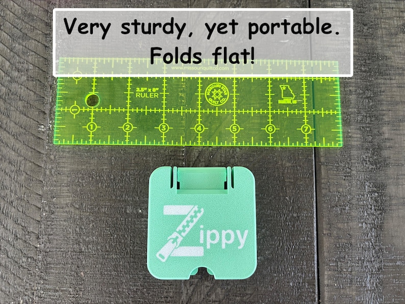 May include: A mint green square folding stand with the white text Zippy and a zipper graphic. A transparent neon green ruler is positioned above the stand, showing a width of approximately 2.5 inches. The text Very sturdy, yet portable. Folds flat! is visible.