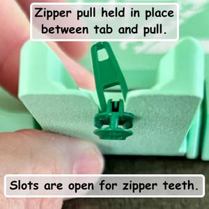May include: A light green plastic zipper repair tool featuring a central slot for zipper teeth and a designated tab to hold the zipper pull in place. The tool includes text labels stating: Zipper pull held in place between tab and pull, and Slots are open for zipper teeth.