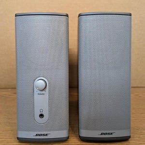 May include: Two gray Bose speakers. The speakers are rectangular with rounded edges and a perforated front. The left speaker has a volume control and headphone jack. The Bose logo is visible on the bottom of each speaker.