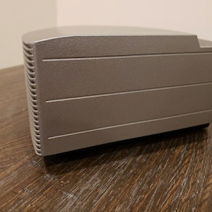 May include: A silver-colored rectangular electronic device with a textured surface and a series of horizontal lines.