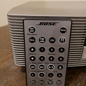 May include: A silver Bose Wave Music System with a remote control. The remote has buttons for On/Off, Sleep, Stop Alarm, Mute, FM/AM, CD, Volume, Presets, Seek/Track, Play/Pause, Stop/Eject, Tune/Playlist, Time, Alarm, Play Mode, Alarm On/Off, Wake to Alarm, and Alarm Time.