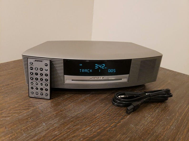 May include: A silver Bose Wave Music System with a digital display showing "3:42" and "TRACK 1 005". The system is on a wooden surface and includes a remote control.