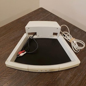 May include: A white, triangular-shaped audio device with a black center panel. It has a rectangular top section and three buttons on the front. Red and white audio cables and a power cord are connected. The device sits on a dark wooden surface.