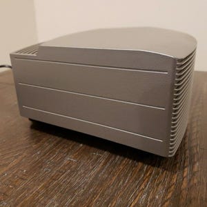 May include: A silver-colored, rectangular-shaped speaker with a slightly curved top and a textured, ribbed side panel.