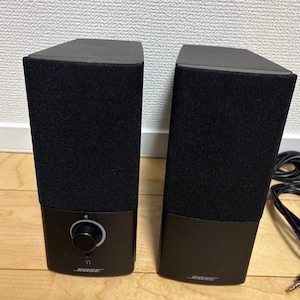 May include: Two black Bose computer speakers. Each speaker has a black fabric front and a black base. The left speaker has a volume control knob and the Bose logo. The right speaker has the Bose logo. Cables are visible.
