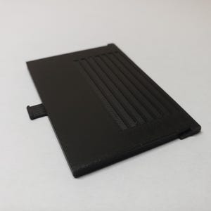 May include: A rectangular, black plastic component with a textured, ridged design on one side. A small tab extends from the left edge. The object appears to be a cover or a part of a larger device.
