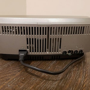 May include: The back of a silver stereo system with a black power cord plugged in. The stereo has a series of vents and several ports on the back.