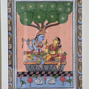 May include: An Indian miniature painting depicting a blue-skinned figure playing a flute, and a woman, seated under a tree. The artwork features intricate details, vibrant colors, and a decorative border, framed in gold.