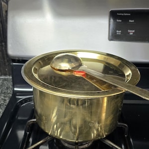 May include: A gold-colored cooking pot with a matching lid and ladle, resting on a black stovetop. The pot is round with a flat bottom, and the ladle rests on the lid. The stovetop has a metal grate and burner.