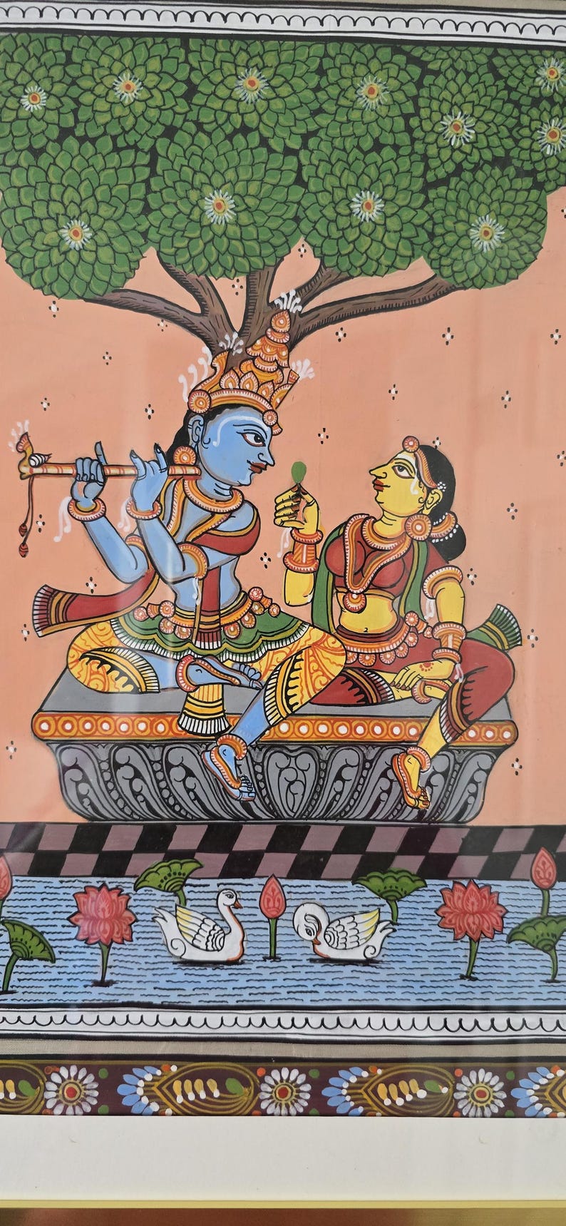 May include: An Indian folk art painting depicting a blue-skinned figure playing a flute, facing a yellow-skinned figure holding a leaf. They sit on a decorative platform under a tree. The scene includes a water feature with swans and lotus flowers.
