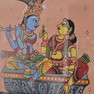 May include: Colorful Indian artwork depicting two figures under a tree. One figure, blue-skinned, plays a flute. The other, yellow-skinned, holds a leaf. The background is peach with a water scene at the bottom.