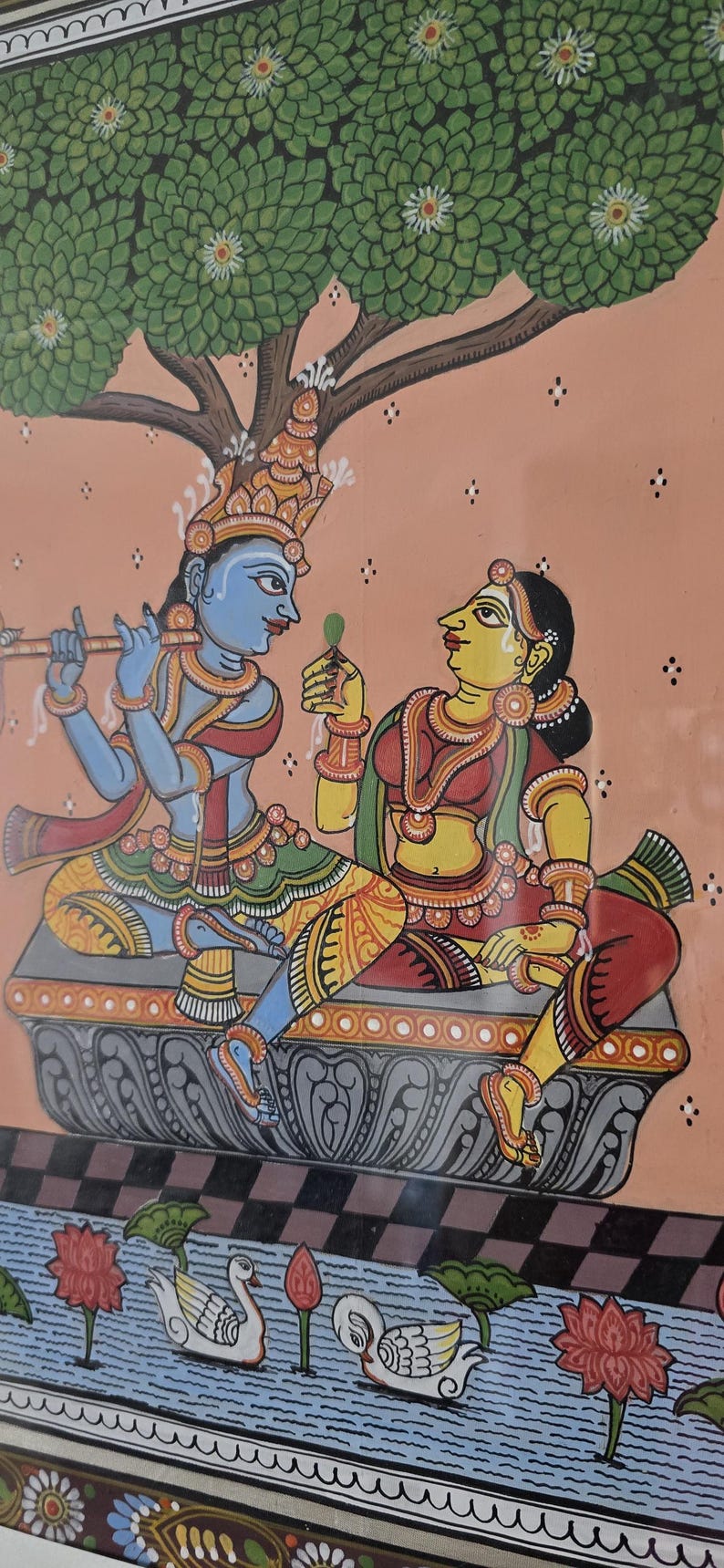 May include: Colorful Indian artwork depicting two figures under a tree. One figure, blue-skinned, plays a flute. The other, yellow-skinned, holds a leaf. The background is peach with a water scene at the bottom.