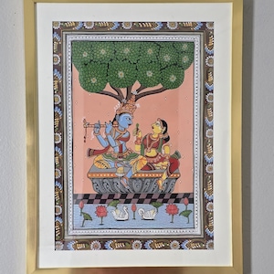 May include: A framed Indian painting depicting a blue-skinned figure playing a flute, seated with a woman under a tree. The artwork features intricate details, vibrant colors, and a decorative border, all encased in a gold frame.