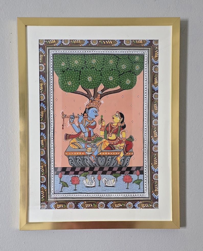 May include: A framed Indian painting depicting a blue-skinned figure playing a flute, seated with a woman under a tree. The artwork features intricate details, vibrant colors, and a decorative border, all encased in a gold frame.