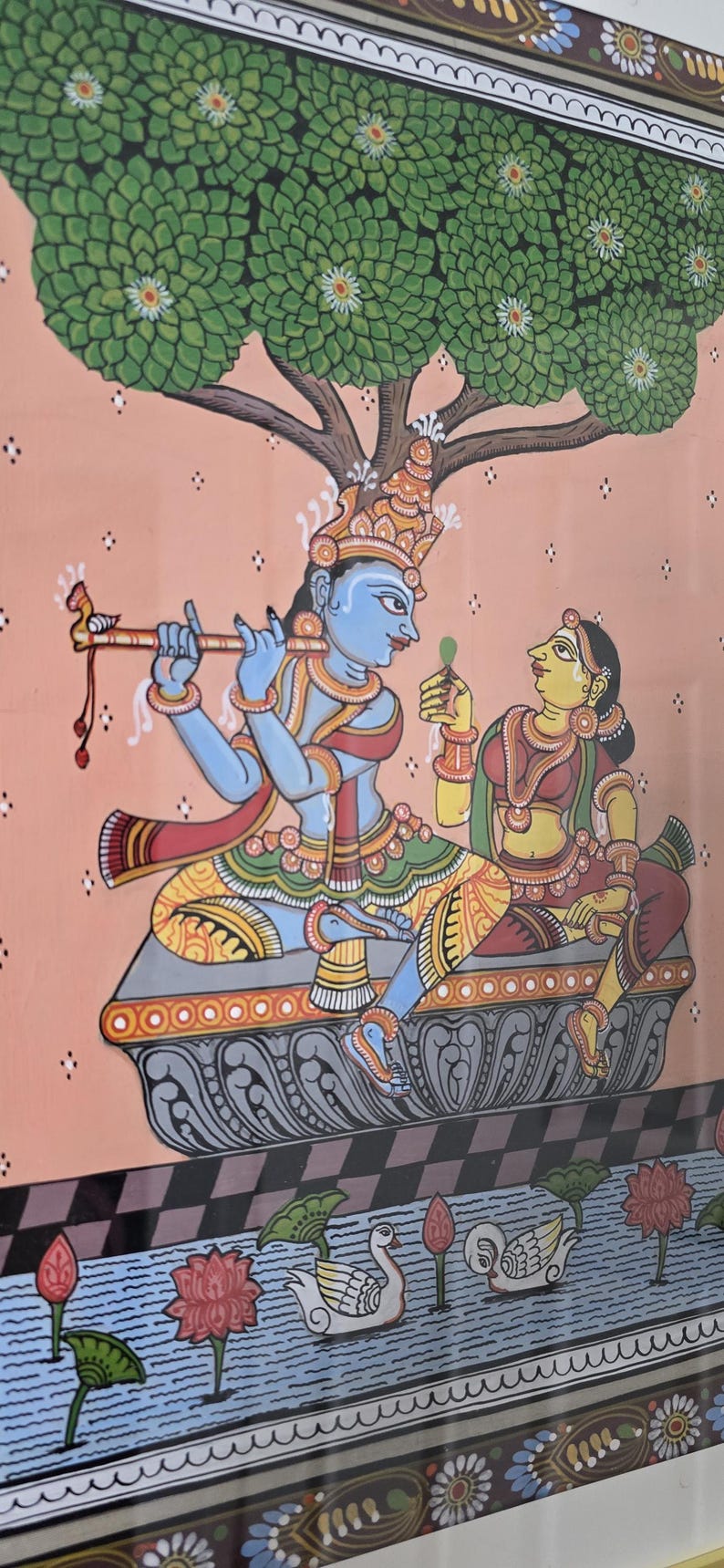 May include: Colorful Indian artwork depicting two figures under a tree. One figure plays a flute, the other holds a leaf. The figures are on a decorative platform above a water scene with swans and flowers. The background is peach.