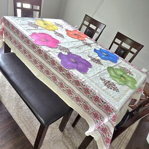May include: A rectangular tablecloth with a floral pattern and large, colorful hibiscus flowers. The tablecloth is cream-colored with a pink and white border. A dark brown bench and chairs surround the table.