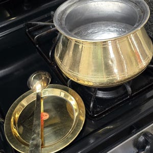 May include: A gold-colored cooking pot with a silver rim sits on a black stovetop. A matching gold plate and serving spoon are next to the pot. The spoon rests on the plate, and a small red and white sticker is visible.