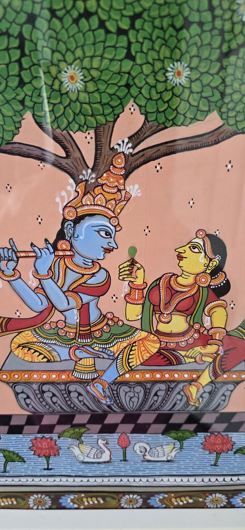 May include: Colorful Indian folk art painting depicting two figures under a tree. The figures are adorned in traditional clothing and jewelry. The background is a soft peach color, and the bottom of the painting shows a pond with swans and lotus flowers.