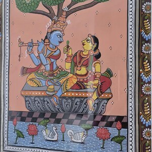 May include: Colorful framed artwork depicting a traditional Indian scene. A blue-skinned figure plays a flute, facing a woman in red and yellow. A tree shades them. The scene includes swans, lotus flowers, and a decorative border.