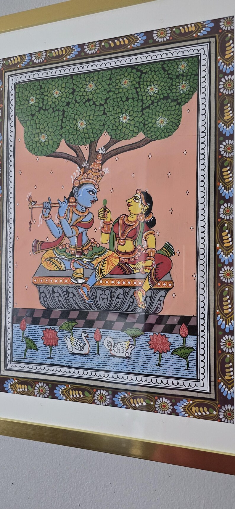 May include: Colorful framed artwork depicting a traditional Indian scene. A blue-skinned figure plays a flute, facing a woman in red and yellow. A tree shades them. The scene includes swans, lotus flowers, and a decorative border.