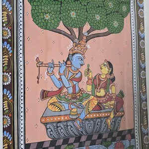 May include: A framed Indian painting depicting a couple under a tree. The figures are in traditional attire, with the male playing a flute. The artwork features vibrant colors, including blue, yellow, and red, and is set against a pink background.